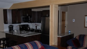 Une cuisine moderne avec des meubles en bois foncé, des appareils électroménagers en acier inoxydable et un îlot central. Le salon est agrémenté d’un canapé bleu à motifs et d’un coussin rouge.