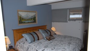 Une chambre à coucher avec une tête de lit en bois, une literie à motifs floraux, une table de chevet avec une lampe, un tableau au mur et une fenêtre avec des stores.