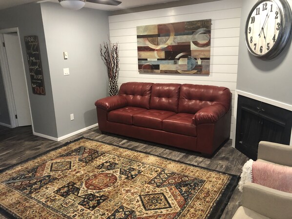 A living room with a brown leather sofa, a patterned rug, a wall clock, and a fireplace.