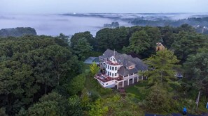 A large, multi-story house surrounded by dense forest, with a view of the sea and mist in the background.