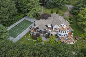 Una casa de varios pisos con cancha de tenis, mobiliario exterior y una bandera.