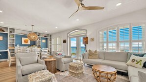 A spacious living room with a grey sofa, two armchairs, a coffee table, and a ceiling fan. There are wicker ottomans, a patterned rug, and a dining area with a white table and chairs. - Seabiscuit -Scenic 30A, Phenomenal Gulf Front Condo w/Spectacular Views! (Rosemary Beach)