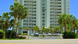 A multi-story hotel with a central vertical feature, surrounded by palm trees and a parking area. - Tidewater 2nd Floor-Awesome Gulf Front 3BR - Indoor & Outdoor Pools (Orange Beach)