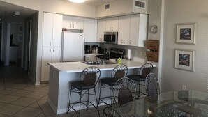 A compact kitchen with white cabinets, a refrigerator, a microwave, and a coffee maker. There are four wicker chairs around a glass table. - Tidewater 2nd Floor-Awesome Gulf Front 3BR - Indoor & Outdoor Pools (Orange Beach)