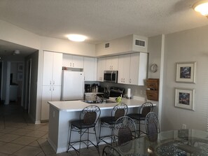 Kitchen area with a central island, refrigerator, microwave, and coffee maker, surrounded by wicker chairs and a glass table.