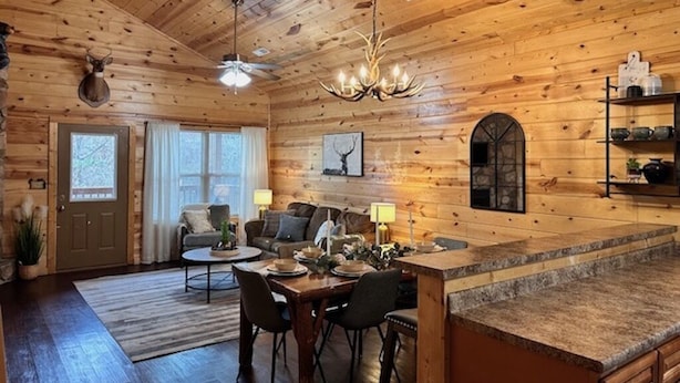 Interior rústico de una cabaña con techo de madera, comedor con mesa y sillas, sofá y zona de cocina con encimera y estantes.