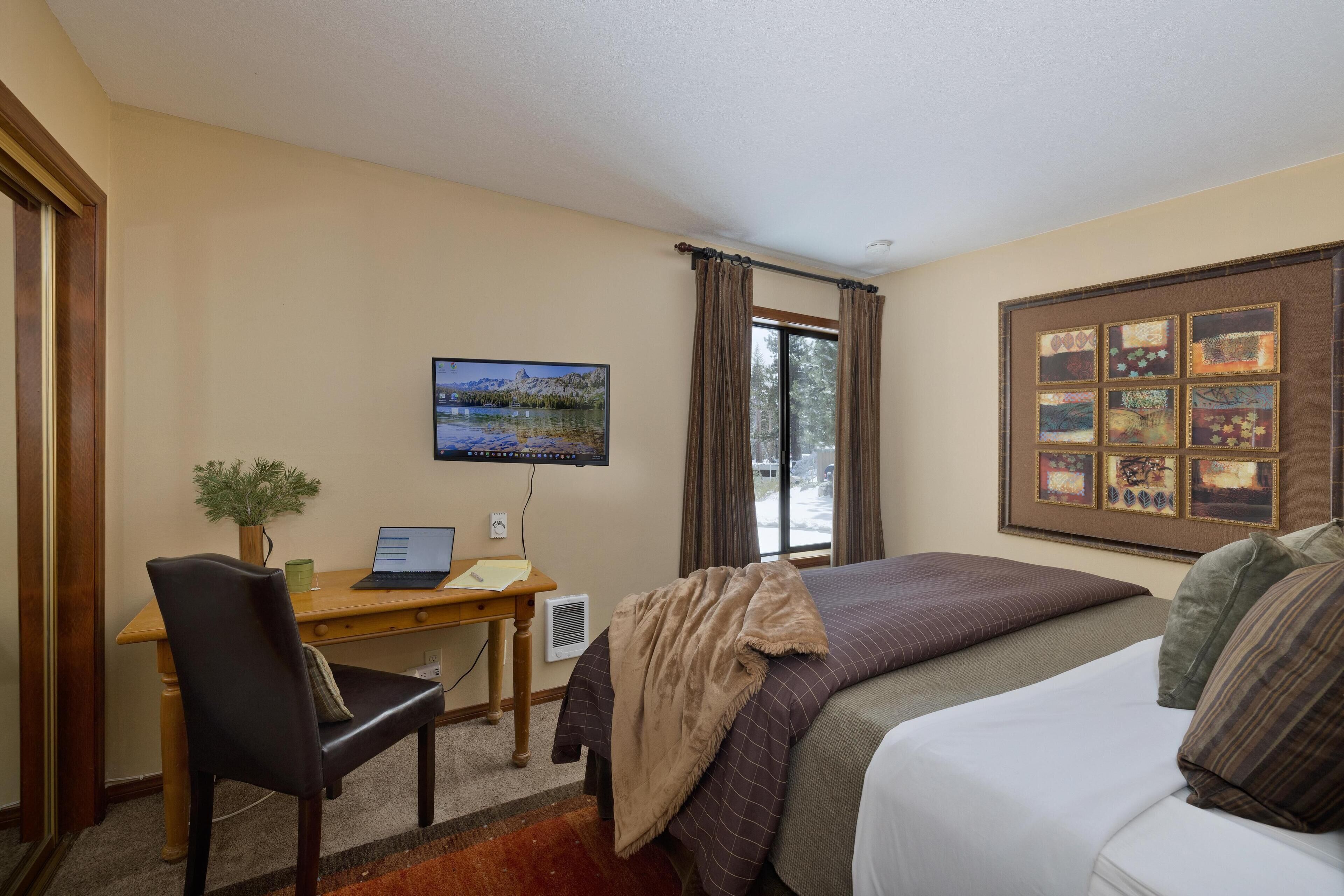 A hotel room with a bed, a desk with a laptop, a chair, a window with curtains, and a wall with framed pictures.