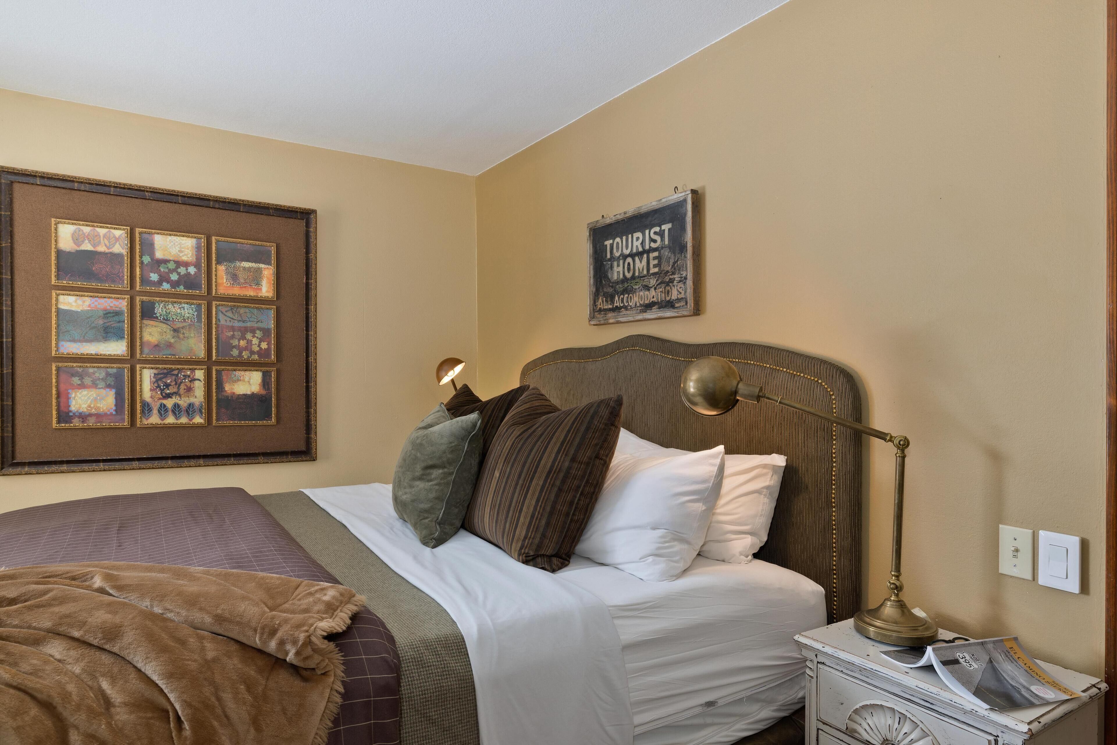 A neatly made bed with a headboard, a bedside lamp, a framed artwork, and a sign on the wall.