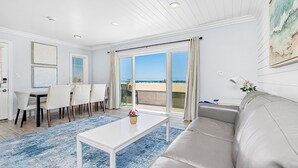 Un salón amplio con un sofá blanco grande, un comedor con sillas, una mesa de centro y vista a la playa a través de un ventanal corredero.