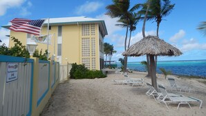 A beachside building with a flag, palm trees, and a thatched umbrella. - Beach Condo in Great Location with Cruzan Rum Bar (Christiansted)