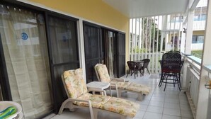 A balcony with two lounge chairs and a small table, a window with a curtain, and a view of a building with balconies. - Beach Condo in Great Location with Cruzan Rum Bar (Christiansted)