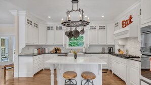 A spacious kitchen with white cabinetry, a large island with stools, a chandelier, and a 'Eat' sign. - The Big Blue House - 6000sf of Luxury in Leiper's Fork (Franklin)