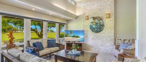A spacious living room with a large TV, comfortable seating, and a view of a lush garden.