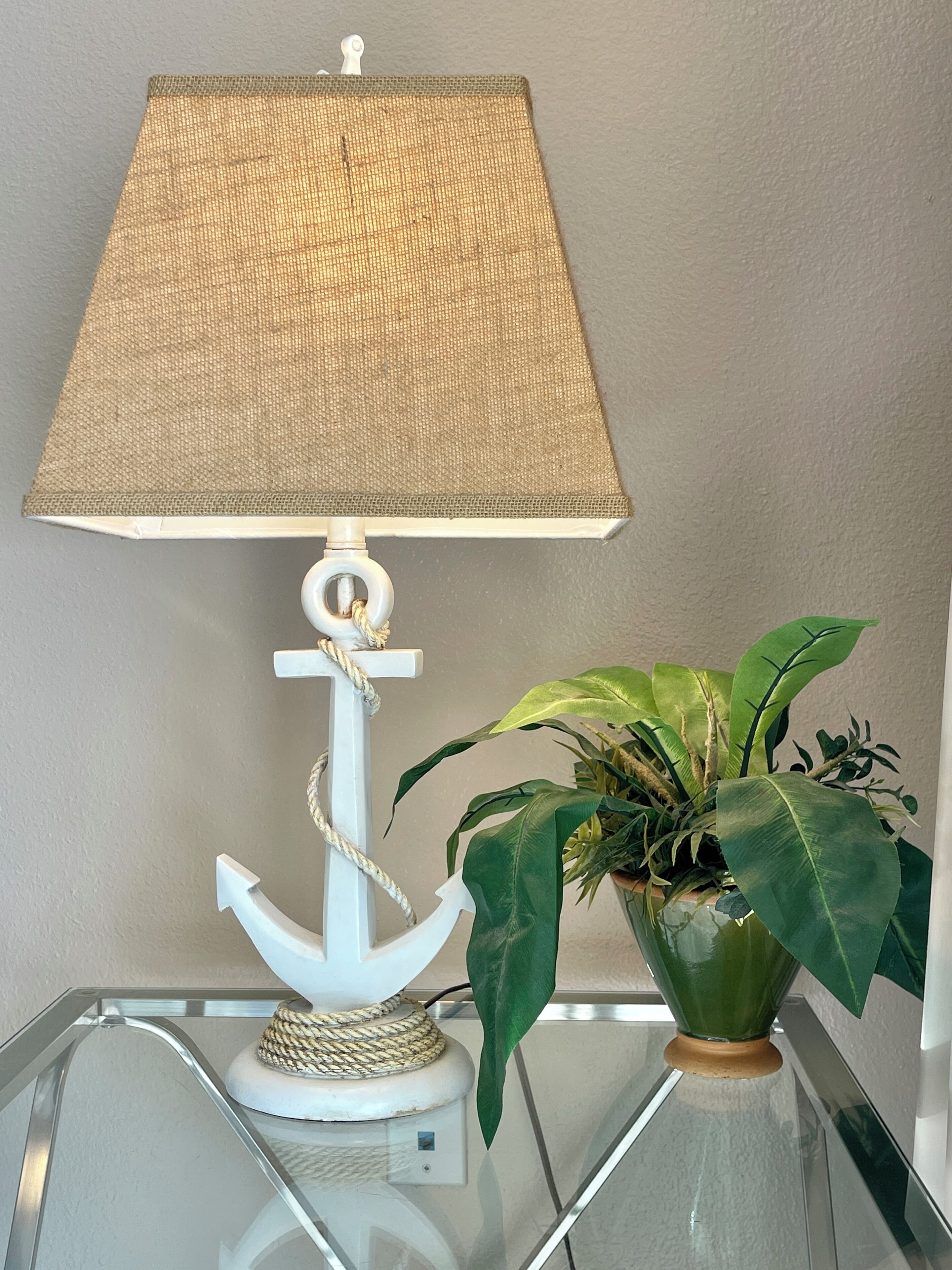 Une lampe avec un pied blanc et un détail en forme de corde, posée sur une table en verre à côté d’une plante en pot.