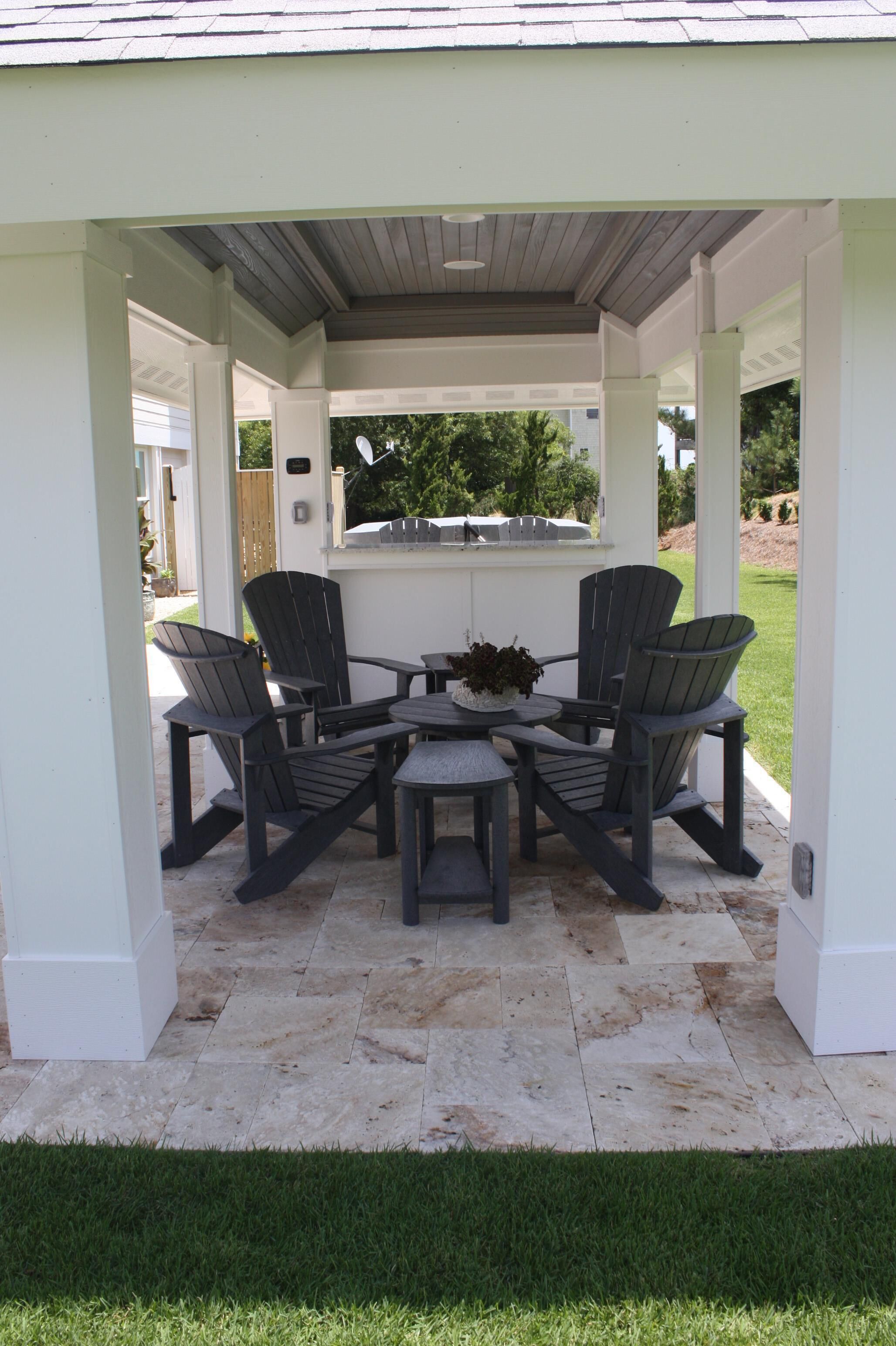 Un patio cubierto con cuatro sillas Adirondack y una mesa pequeña, rodeado de pilares blancos y un suelo de baldosas.