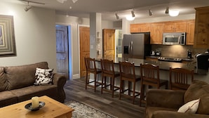 Un soggiorno con un divano in pelle marrone, un tavolino in legno e un tappeto decorato. È presente una zona cucina con mobili in legno, un frigorifero e un forno a microonde.