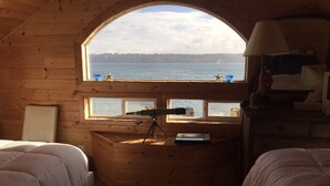 A cozy cabin with a large arched window overlooking a body of water, two beds, a telescope, and a lamp on a nightstand. - Anchor Away, Whidbey Island Beachfront Getaway (Clinton)
