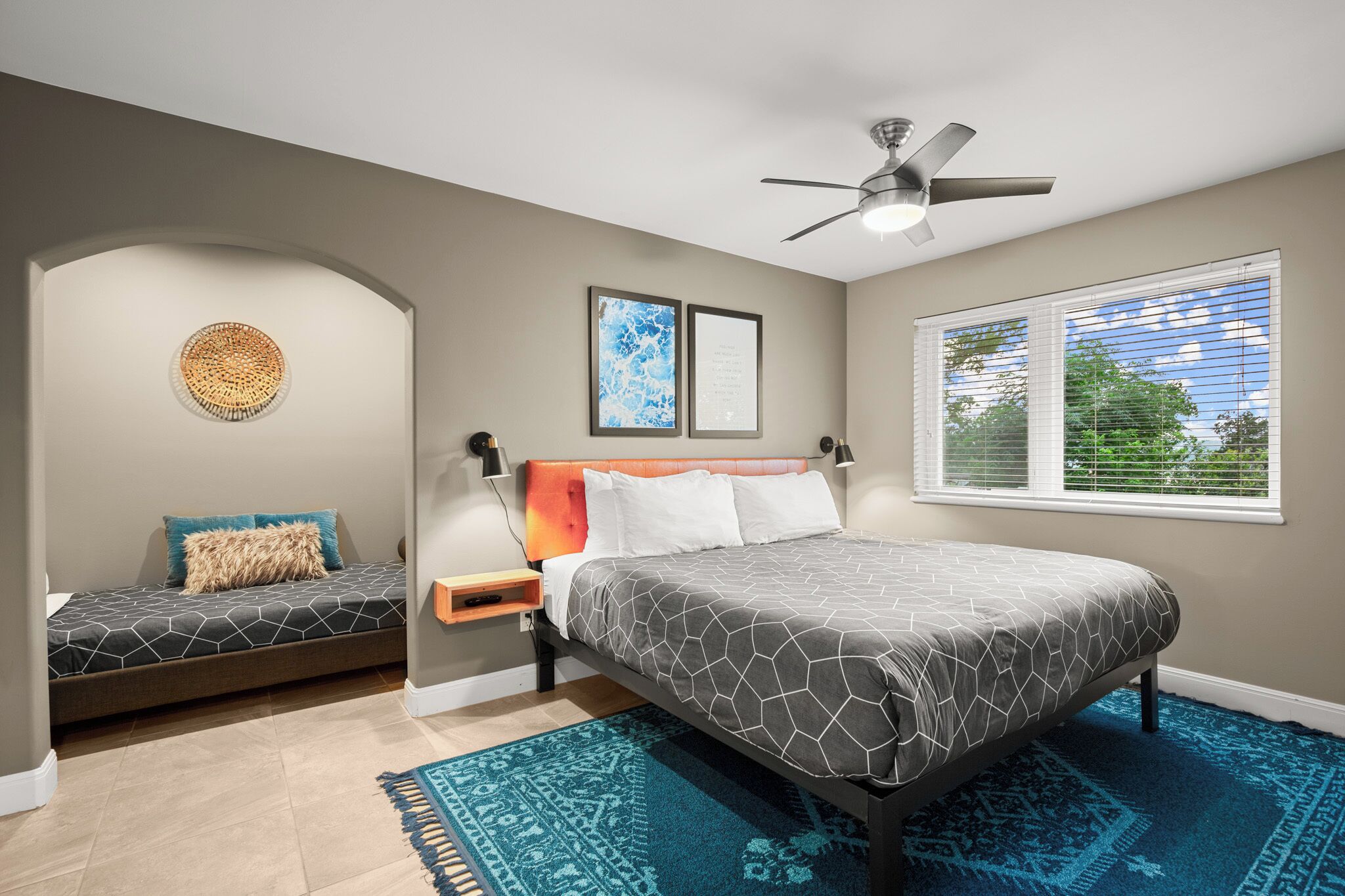 A bedroom with a bed, a smaller bed, a ceiling fan, a window with blinds, and a wall-mounted artwork.