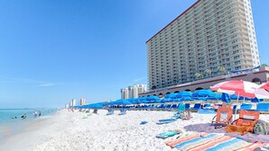 A beach with blue umbrellas and lounge chairs, a tall hotel building, and a clear blue sky.