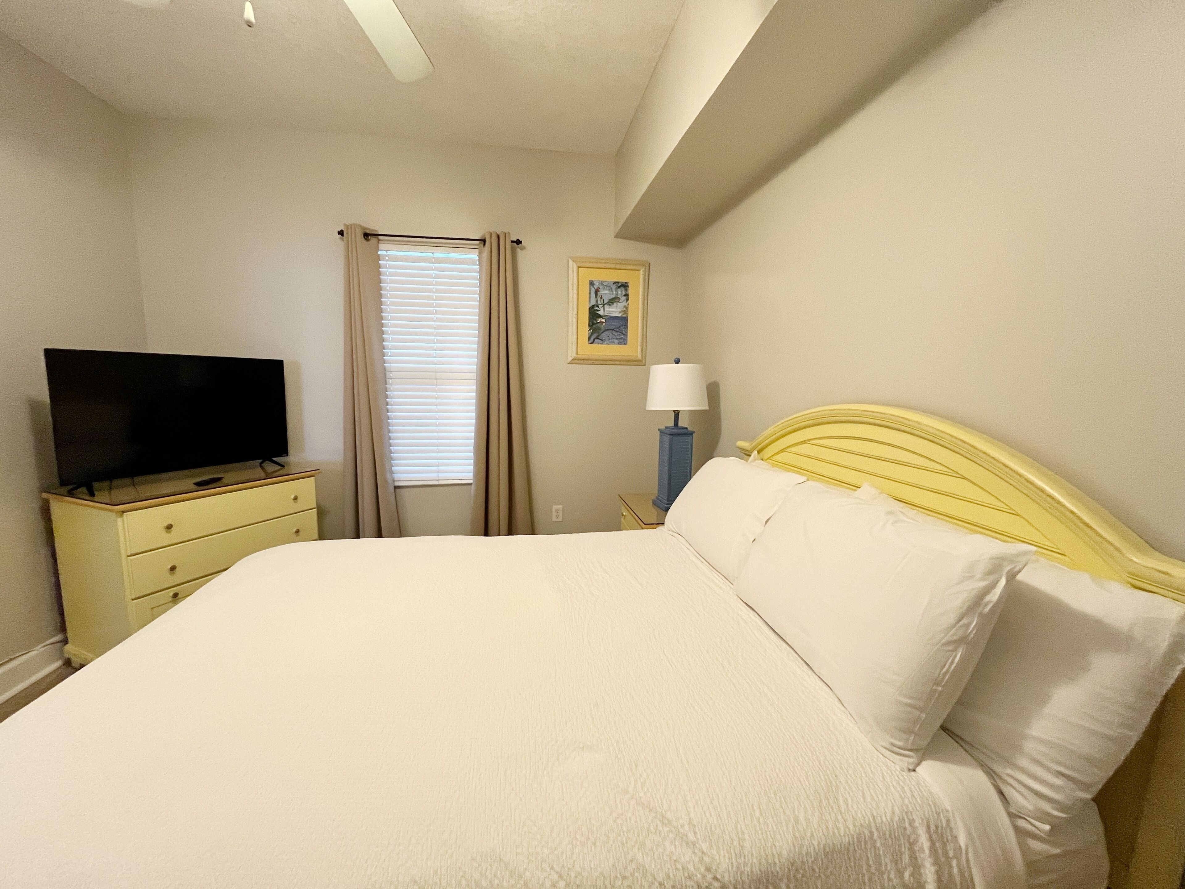 Un dormitorio con cama, televisor en una cómoda, lámpara y ventana con cortinas.