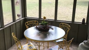 A covered patio with a round table and yellow metal chairs, a potted plant, and a view of a grassy area with trees and a building in the distance. - ACE OF HEARTS The perfect getaway! Only 3.5 miles from Main Street. (Fredericksburg)