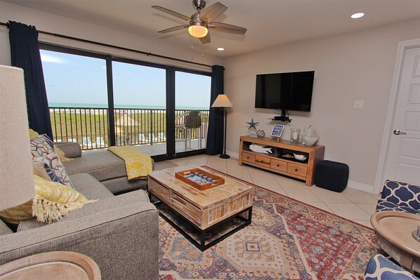 A living room with a sofa, a coffee table, a TV, and a view of the beach.