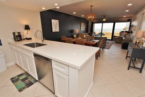 A modern kitchen with a central island, a dining area with wicker furniture, and a view of the sea through large windows.