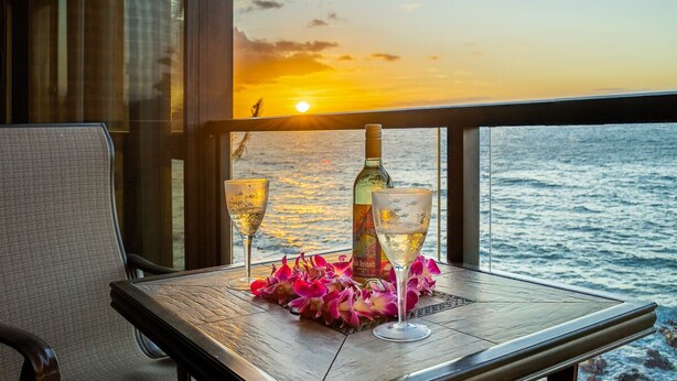A table with two glasses of white wine, a bottle, and pink flowers on a balcony overlooking the ocean at sunset. - 402 - Spectacular Oceanview Unit with A/C! 2 sofa beds! (Kihei)