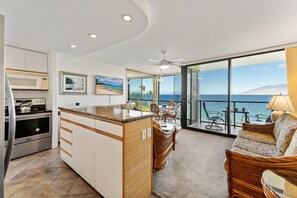 A modern kitchen with a central island, dining area with wicker furniture, and a balcony with a view of the ocean.