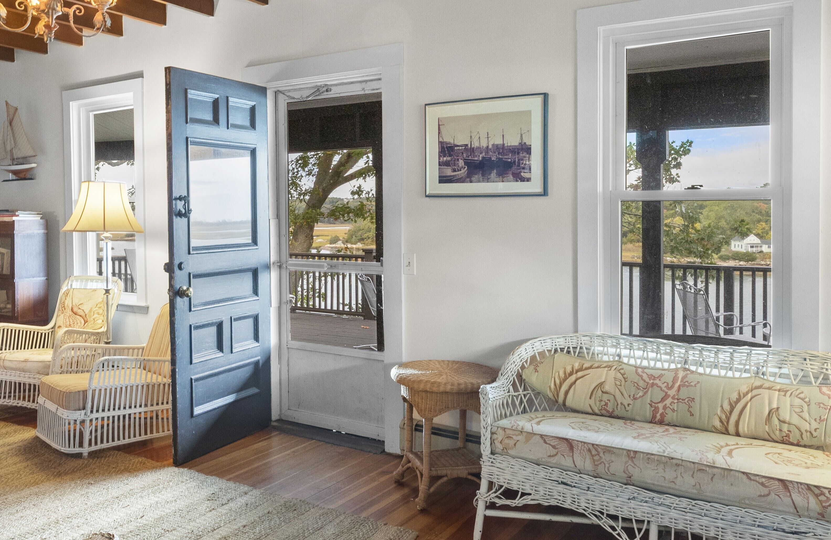 Habitación acogedora con un sofá de mimbre, una silla blanca y una mesita. Una puerta azul da a un área exterior con barandilla.