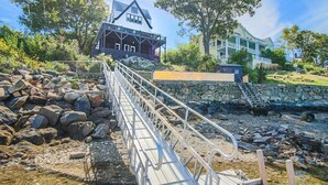 Eine Holzbrücke führt zu einem Haus mit Balkon, umgeben von Felsen und Bäumen.