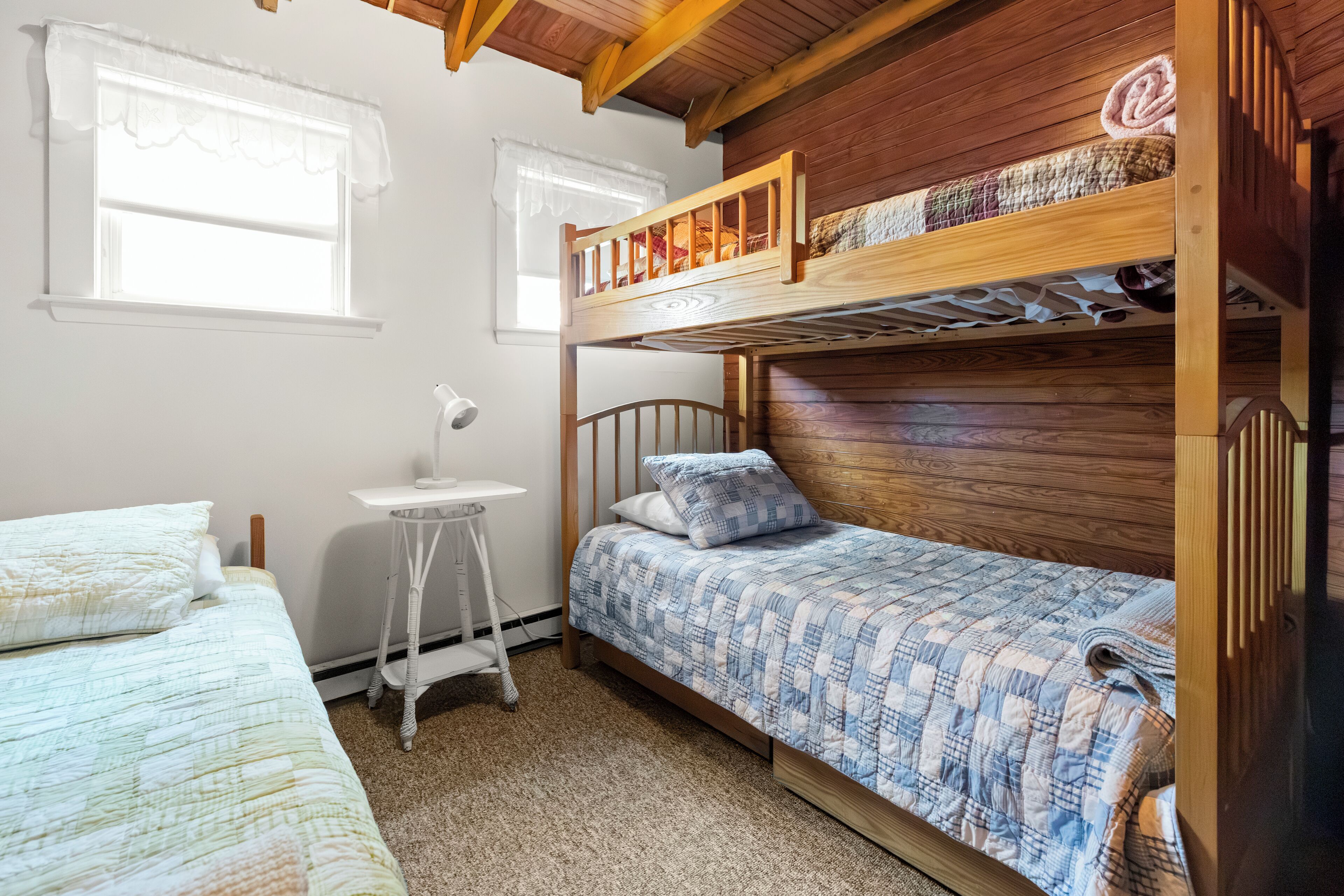 Quarto com beliche, cabeceira de madeira, pequena escrivaninha e janela com cortina branca.