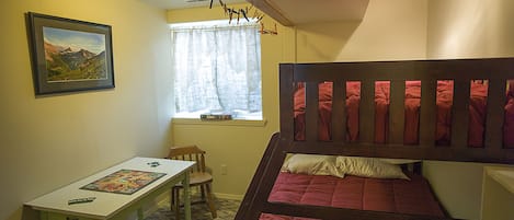 A room with a bunk bed, a desk with a board game, a chair, a window with curtains, and a framed picture on the wall.