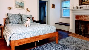 Ein Schlafzimmer mit einem hölzernen Bett, gemusterten Betttextilien, einem kleinen Hund, einem Kamin und einer Tür zu einem anderen Raum.