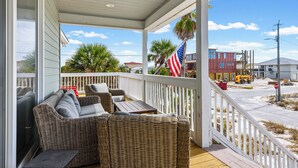 A covered porch with wicker furniture and an American flag. - 🌞 The Sunshine Gulfside | Sleeps 22 w/ Private Pool + Steps to Beach (Pensacola Beach)