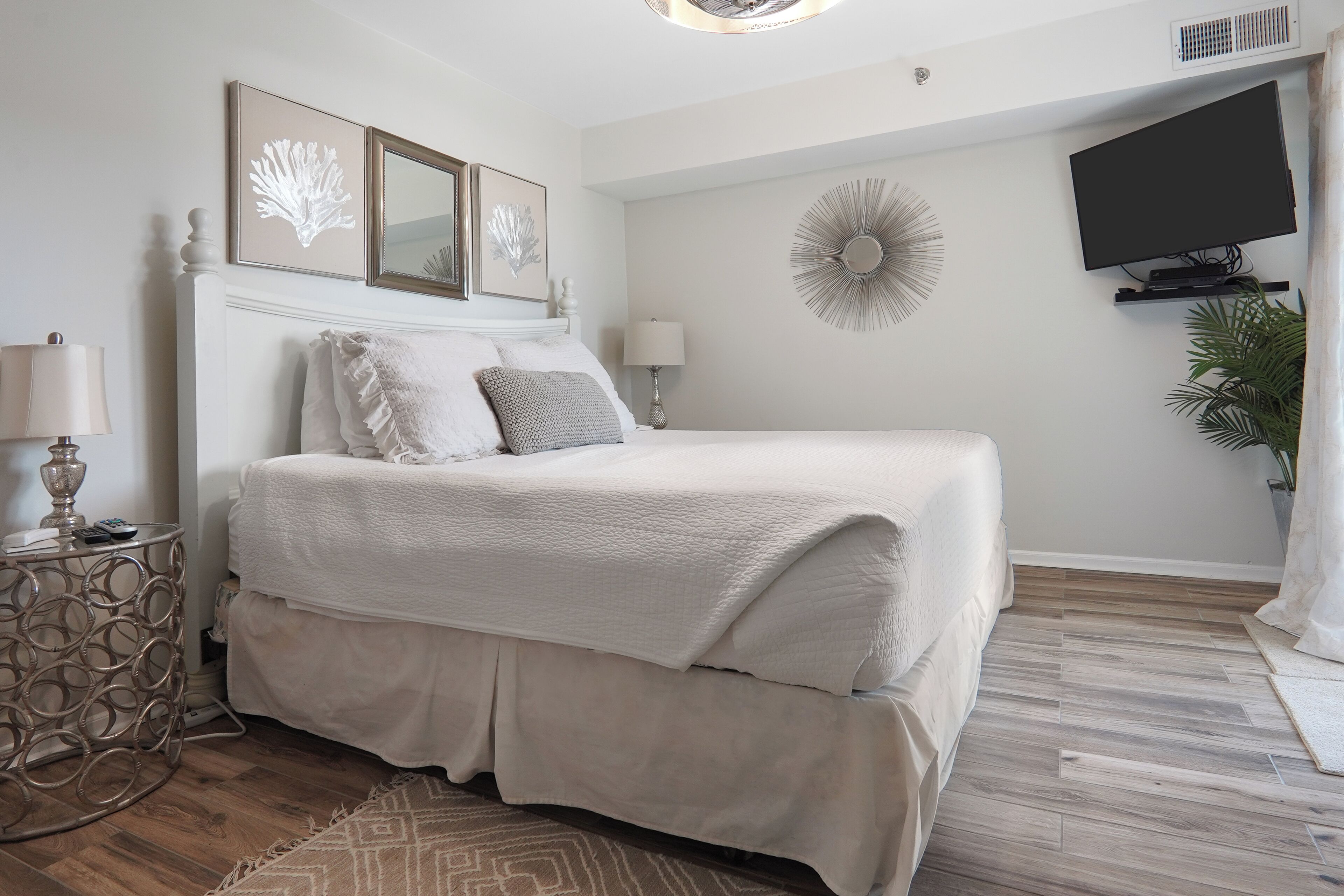 A bedroom with a large bed, a side table with a lamp, a wall-mounted TV, and a decorative mirror.