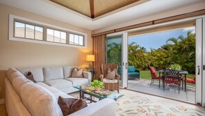 A living room with a sofa, armchairs, a glass coffee table, and a view of a tropical garden through a sliding glass door.