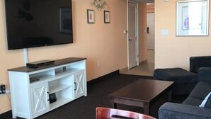 A living room with a flat-screen TV, a white entertainment unit, a dark coffee table, and a gray sofa. - Oceanfront Penthouse Suite - Prince Resort (Myrtle Beach)