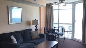 A hotel room with a dark-colored sofa, a small table, a lamp, and a view of the balcony through large windows. - Oceanfront Penthouse Suite - Prince Resort (Myrtle Beach)
