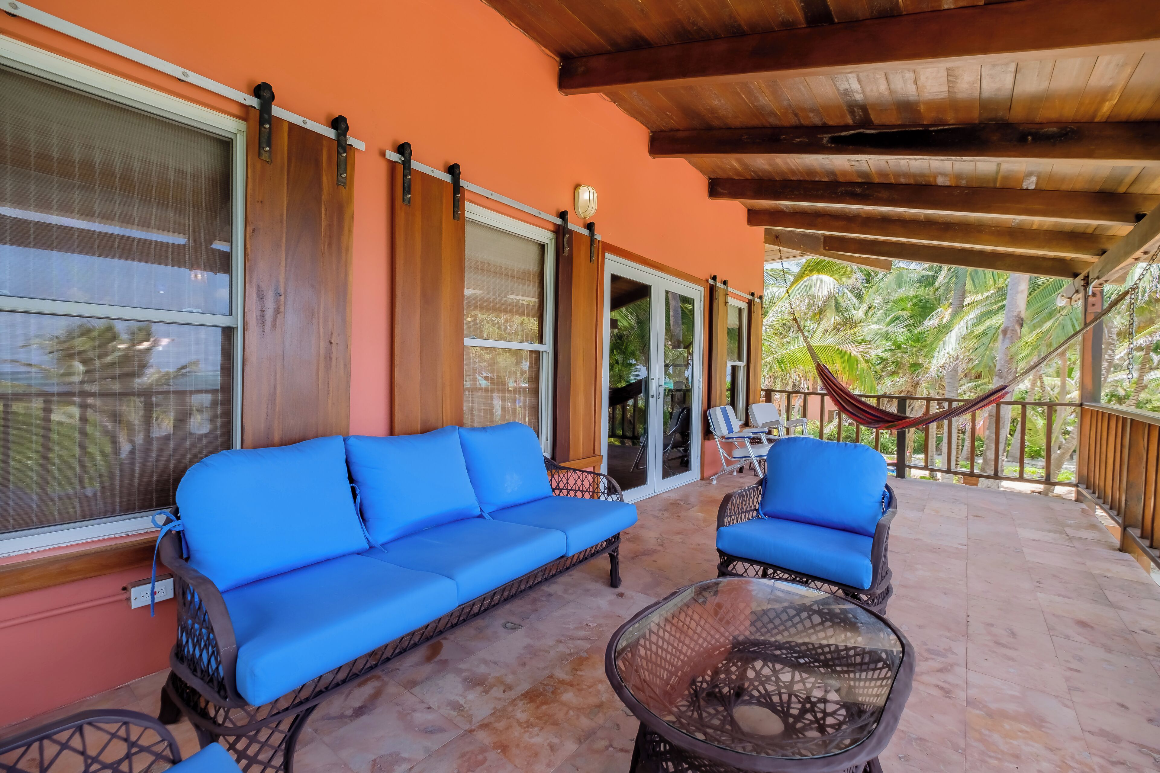 Une terrasse couverte avec des canapés bleus dotés de coussins et une table en verre, donnant sur un jardin tropical.