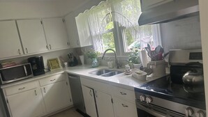 Une cuisine avec des meubles blancs, un évier en acier inoxydable, une cuisinière et une fenêtre ornée de rideaux en dentelle.