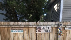 Two green Adirondack chairs facing each other with a small table in between, set against a wooden fence with signs reading "BEACH HOUSE" and "AT THE SHORE". - Family Friendly Townhome W/POOL In Ocean City, Md. (Ocean City)