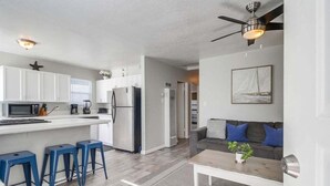 A modern kitchen with white cabinets, a refrigerator, and a microwave. There are three blue bar stools and a wooden coffee table with a plant. - Large 5 Bedroom House Beach side in Mission Beach (San Diego)