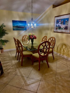 Un comedor con una mesa de vidrio rodeada de sillas de madera, una lámpara colgante y dos cuadros en las paredes.