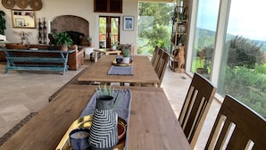 Een ruime woonkamer met een houten eettafel, zachte zithoeken, een open haard en een groot raam met uitzicht op de omgeving.