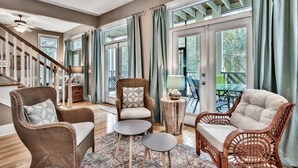A spacious living area with wicker furniture, a staircase, and large windows with curtains.