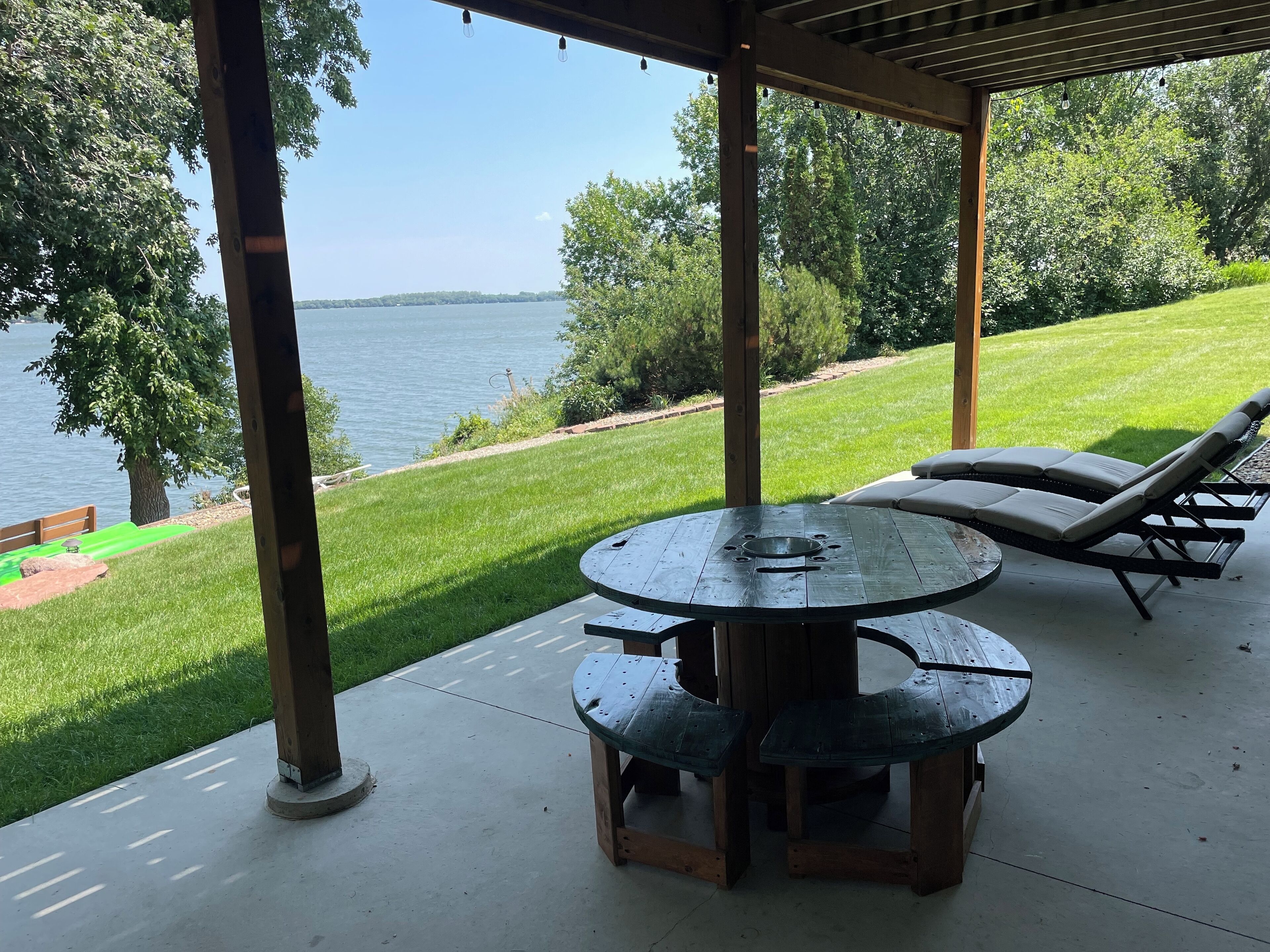 A covered patio with a round table and two lounge chairs, overlooking a grassy area and a body of water.