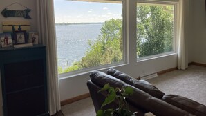 A room with a large window overlooking a body of water, a brown leather sofa, and a blue cabinet with framed pictures. - Beautiful home on Clear Lake! Amazing views 🌅 Great fishing 🎣 Great boating 🚤 (Lake City)