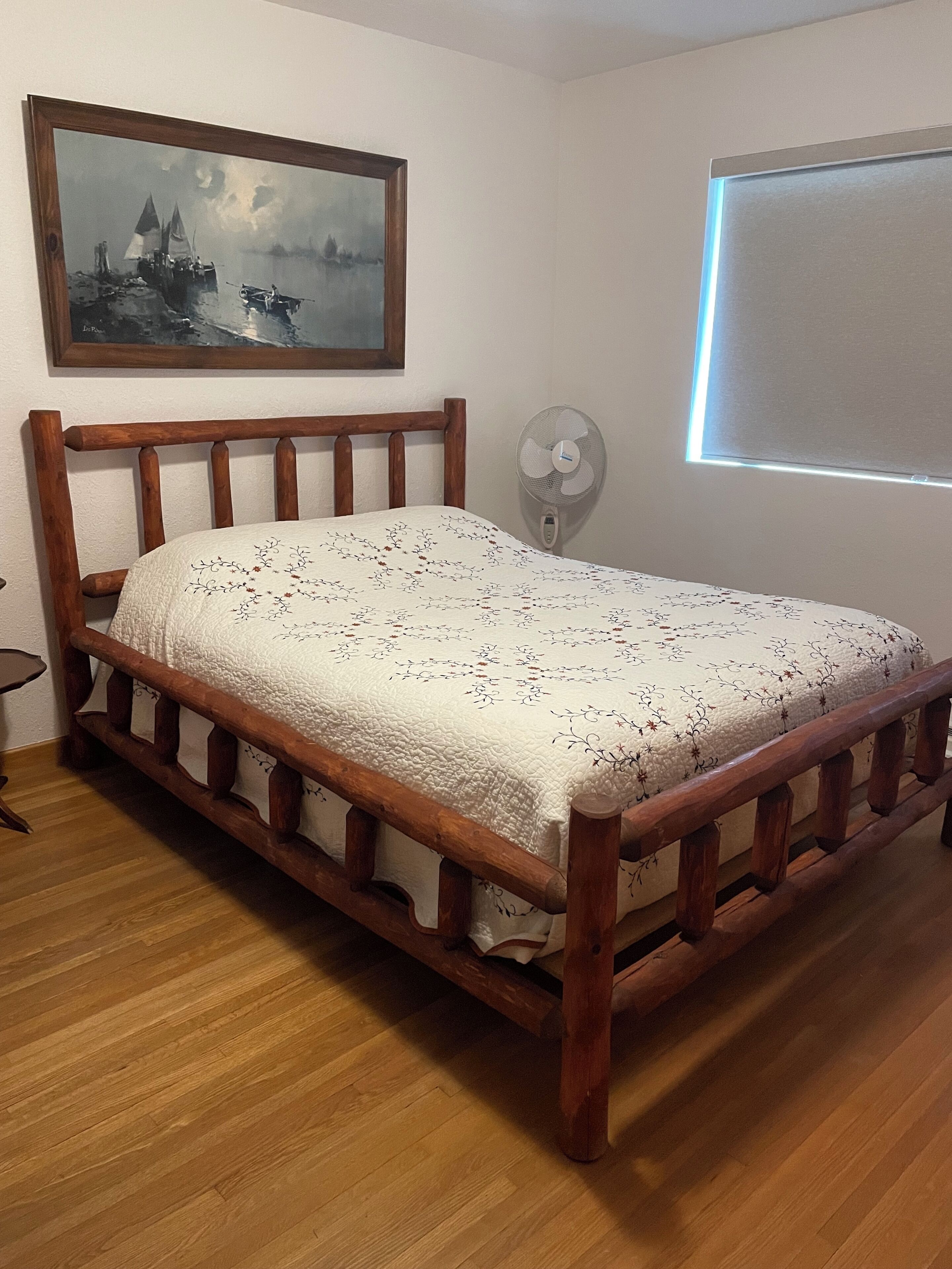 A wooden bed with a floral bedspread in a room with a wooden floor, a wall-mounted fan, and a painting above the bed.