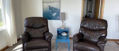 Een kamer met twee bruine leren fauteuils, een blauwe bijzettafel met lamp en klok, en een groot abstract schilderij aan de muur.
